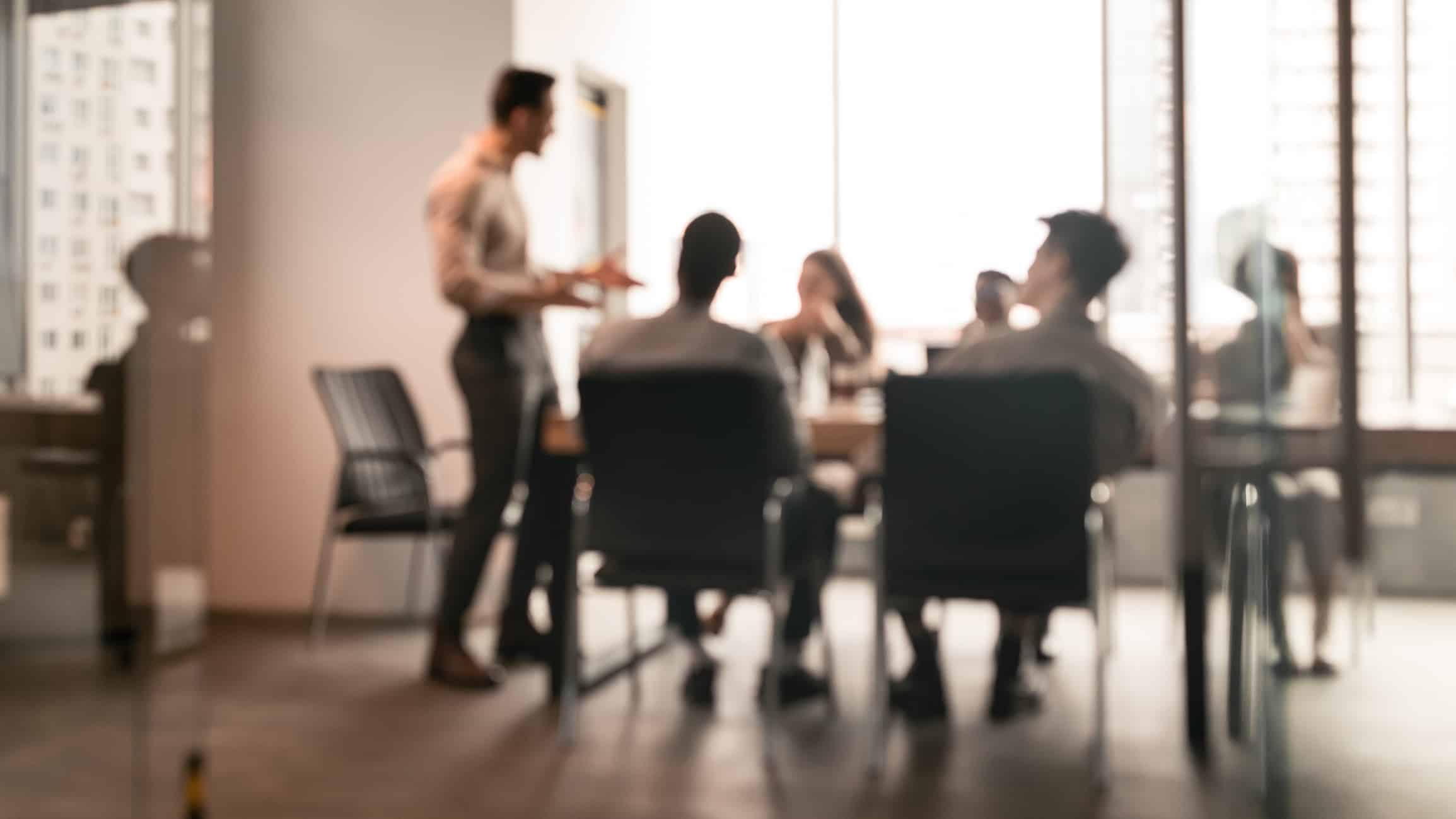 Colleagues having meeting in boardroom, businessman giving speech, blurred photo. ISO 44001 promotes collaborative business relationships.